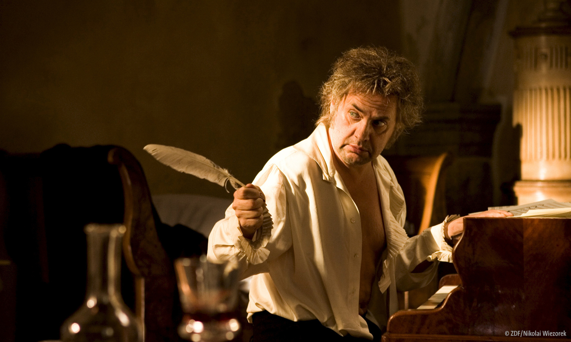"Giganten: Beethoven - Genie am Abgrund": Spielszene: Beethoven (Uwe Ochsenknecht) sitzt an einem Klavier. Er hält eine Schreibfeder in der linken Hand und schaut zur Seite. © ZDF/Nikolai Wiezorek