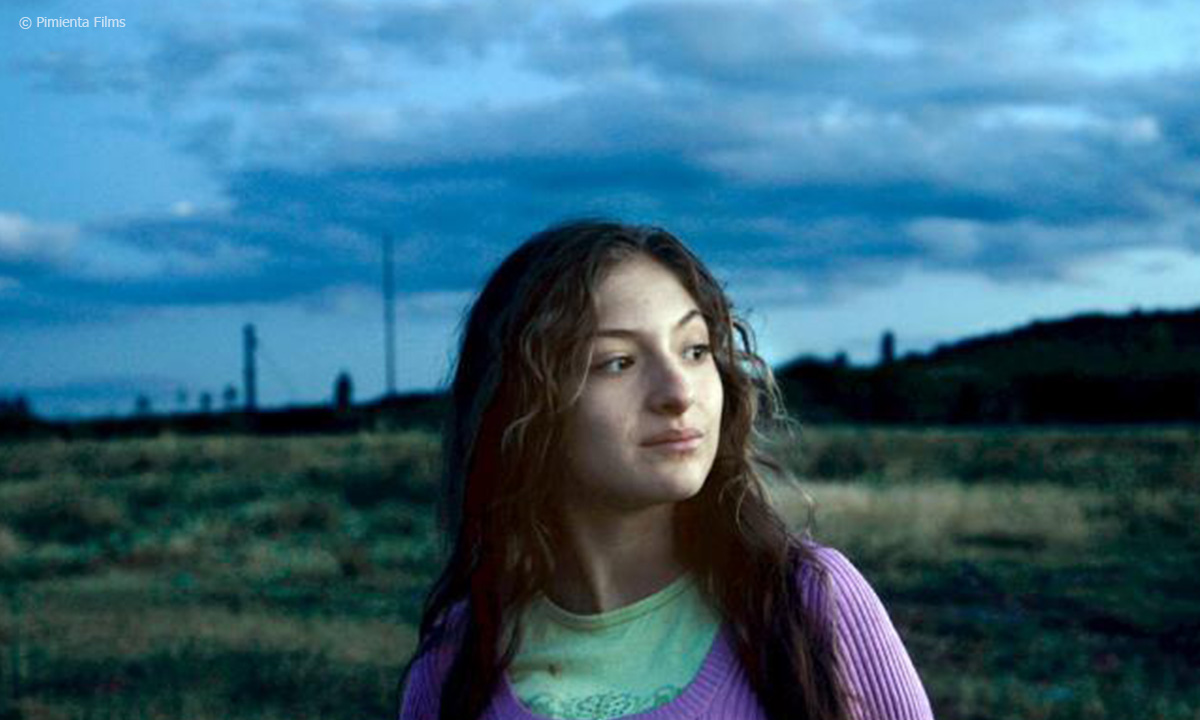 Filmstill aus "Tempestadt". Zu sehen ist eine junge Frau, die im Freien steht. Der Himmel ist dunkel und wolkenverhangen.